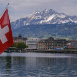 A swiss flag over the water with a city in the background